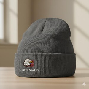 Może przedstawiać: Szara czapka beanie z wywiniętym brzegiem. Czapka posiada haftowany wzór orła i małej amerykańskiej flagi. Słowa "UNITED STATES" są wyhaftowane poniżej. Czapka wykonana jest z prążkowanej dzianiny.