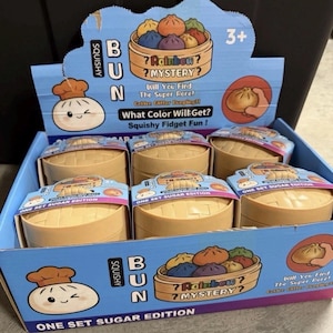 May include: A display box of "Squishy Bun" mystery toys. The box is blue with cartoon illustrations of buns and text that reads "Rainbow Mystery". Each individual toy is in a tan container with a lid. The box is labeled "One Set Sugar Edition".
