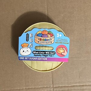 May include: A yellow plastic container with a blue and pink label featuring a cartoon dumpling and the text "BUN". The label also says "Rainbow Mystery" and "Squishy Fidget Fun!"