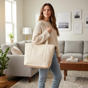 May include: A cream-colored tote bag with sturdy handles is held by a person. The bag is rectangular with a simple design, and the person is wearing a cream sweater and blue jeans. The background includes a living room setting with a couch and artwork.