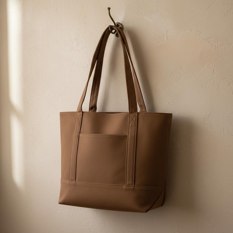 May include: A brown tote bag with long handles hangs from a hook on a beige wall. The bag has a rectangular shape with a front pocket and reinforced stitching details. The bag is made of a sturdy material.