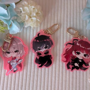 May include: Three acrylic keychains featuring chibi-style anime characters with pink outlines and gold-toned lobster clasps. The left character has light hair, the centre character has black hair, and the right character has long red hair.