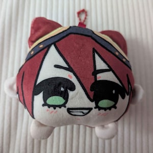 May include: A plush toy featuring a character with short red hair, a dark headband with yellow accents, and large green eyes. The character has a pale face with a small smiling mouth and pink blush marks on the cheeks. A small red ball chain is attached to the top.