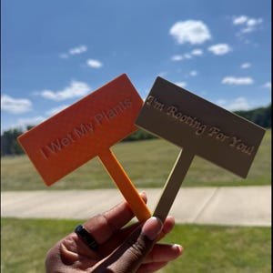 May include: Two rectangular plant markers with stakes, one orange with the text I Wet My Plants and one tan with the text I'm Rooting For You!. The markers have a flat surface and a pointed stake at the bottom for insertion into soil or pots.