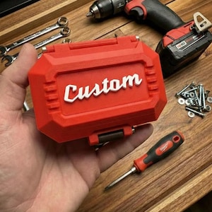 May include: Red "Custom" tool case with a red and black drill, screwdriver, and other tools. The case has the word "Custom" in white lettering. The tools and case are on a wooden surface.