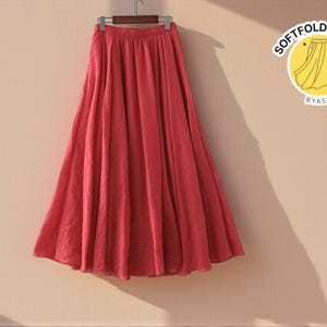 May include: A flowing, coral-colored skirt hangs from a wooden hanger. The skirt is gathered at the waist and flares out to a full, floor-length hem. The fabric appears lightweight and slightly textured. A logo with the text "SOFTFOLSTUDIO BYASH" is visible.