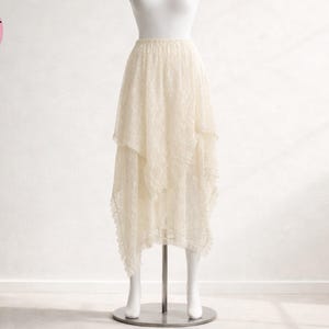 May include: Ivory lace skirt with an asymmetrical hemline, displayed on a mannequin. The skirt has a tiered design and a delicate, flowing appearance. The text "SKIRT STYLE" is in a pink circle in the upper left corner.