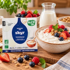 Pode incluir: Um pacote de preparação para skyr da marca Nat-ali com um gráfico circular azul, texto branco e um logótipo de certificação orgânica. O pacote está ao lado de uma tigela de cerâmica cheia de skyr branco com morangos, mirtilos e granola.