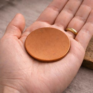 Può includere: Un piccolo disco circolare e piatto con una superficie liscia e opaca color rame. L'oggetto è solido e di colore uniforme, caratterizzato da una forma geometrica semplice con bordi netti e definiti, appoggiato su un palmo.