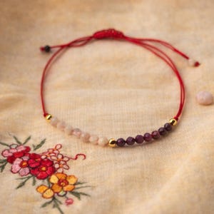 Pode incluir: Pulseira de contas com cordão vermelho. Apresenta um gradiente de contas rosa e roxas, acentuadas com contas douradas. A pulseira é exibida em tecido amarelo com bordados florais.