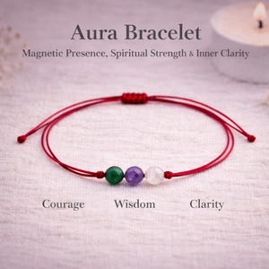 Op de afbeelding: Aura Armband met een rood koord en drie edelsteen kralen: groen, paars en wit. De armband is gelabeld met de woorden "Moed", "Wijsheid" en "Helderheid". De tekst "Magnetische aanwezigheid, spirituele kracht & innerlijke helderheid" is ook zichtbaar.