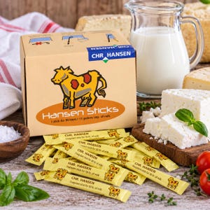 Op de afbeelding: Een kartonnen doos met het label Chr. Hansen Hansen Sticks met een gele koe-illustratie, met daarin meerdere gele individuele staafjes voor het maken van kaas, elk gemarkeerd voor 50 liter melk, weergegeven met kaas en melk.