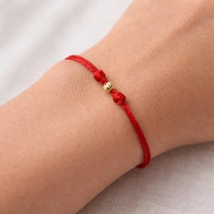 Op de afbeelding: Een rode draadarmband met een gouden kraal in het midden. De armband is aan weerszijden van de kraal met knopen vastgemaakt. De armband wordt om een pols gedragen.