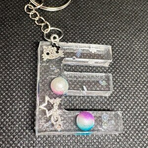 May include: A clear resin keychain in the shape of the letter "E". The keychain is adorned with a silver charm, iridescent beads, and two spherical beads with pink, purple, and blue gradients. A silver keyring and chain are attached.