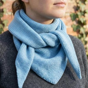 Puede incluir: Bufanda de punto azul claro anudada en la parte delantera. La bufanda está hecha de un material suave y texturizado y es de color azul claro liso. La persona lleva un jersey gris.