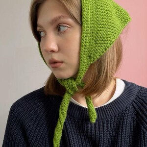 Puede incluir: Un pañuelo de cabeza tejido de color verde brillante atado bajo la barbilla. El cubrecabezas tiene forma cuadrada y está hecho de hilo texturizado. La persona lleva un jersey azul marino.