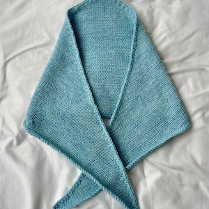 May include: A light blue knitted scarf with a triangular shape. The scarf is made of soft yarn and has a textured appearance. The scarf is displayed on a white surface.