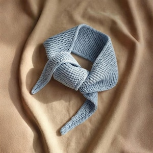 Puede incluir: Bufanda de punto azul claro, anudada en el centro. La bufanda está hecha de un material suave y texturizado y se muestra sobre un fondo de tela beige. Los extremos de la bufanda son puntiagudos.