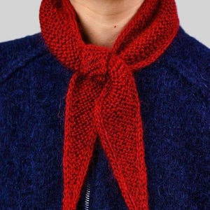 May include: A close-up of a red knitted scarf tied around a person's neck. The scarf is a vibrant red colour and has a textured appearance. The person is wearing a dark blue jacket with a silver zip.
