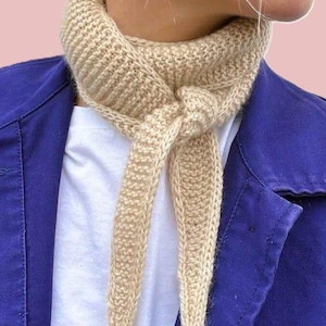May include: A beige knitted scarf tied in a knot. The scarf is made of a thick yarn and has a textured appearance. The scarf is worn over a white shirt and a blue jacket. The background is a light pink color.