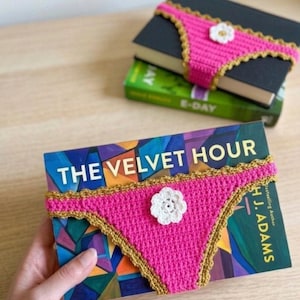 Puede incluir: Libros con una funda de libro de ganchillo rosa con forma de braga. La funda tiene un ribete dorado y una flor blanca. El libro se titula "The Velvet Hour".