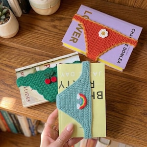 Könnte beinhalten: Gehäkelte Buchaccessoires in Form von Badebekleidung. Eines ist orange mit einer weißen Blume, ein anderes ist grün mit Kirschen und das dritte ist blau mit einem Regenbogen. Die Accessoires befinden sich auf Büchern.