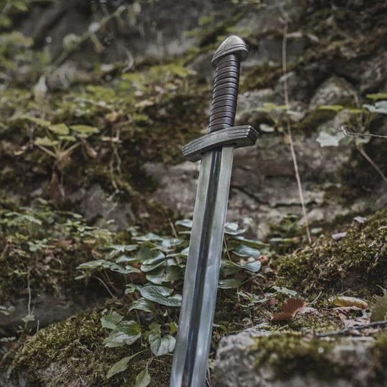 May include: A medieval sword with a silver blade and a dark brown, textured handle. The sword is positioned upright against a backdrop of moss-covered rocks and foliage, creating a rustic aesthetic.