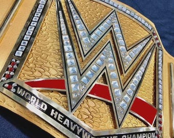 May include: A gold-colored championship belt with the letters "WWE" in the center, encrusted with clear and red gemstones. The words "WORLD HEAVYWEIGHT CHAMPION" are displayed in black lettering on the belt.