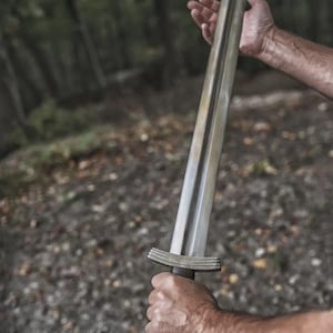 May include: A silver-colored sword with a wide, flat blade and a dark brown handle. The sword is held in a person's hands against a blurred forest background. The hilt features a crossguard and a rounded pommel.