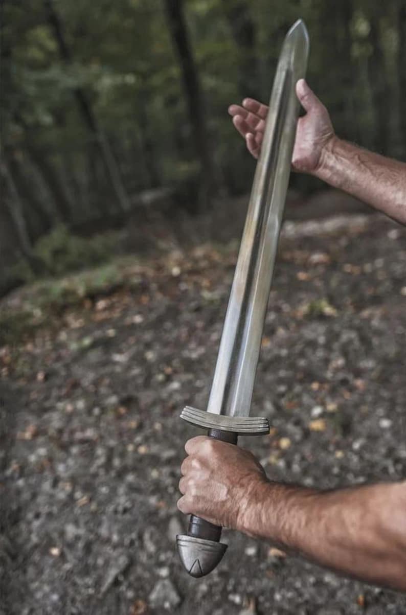 May include: A silver-colored sword with a wide, flat blade and a dark brown handle. The sword is held in a person's hands against a blurred forest background. The hilt features a crossguard and a rounded pommel.