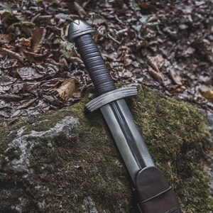 May include: A Viking-style sword with a silver blade and guard, a dark leather-wrapped handle, and a rounded pommel. The sword rests on a moss-covered rock, with fallen leaves in the background.