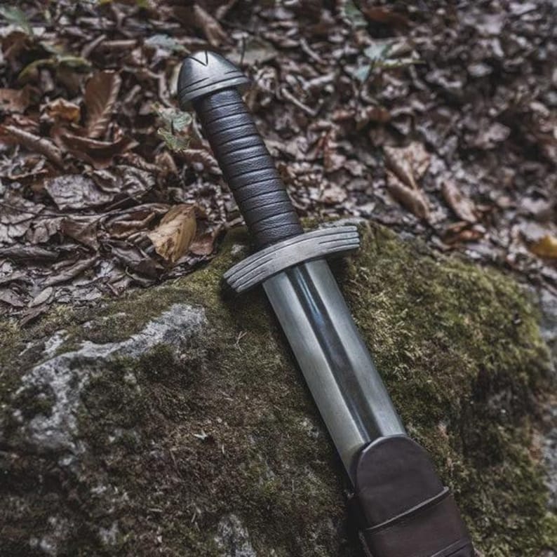 May include: A Viking-style sword with a silver blade and guard, a dark leather-wrapped handle, and a rounded pommel. The sword rests on a moss-covered rock, with fallen leaves in the background.