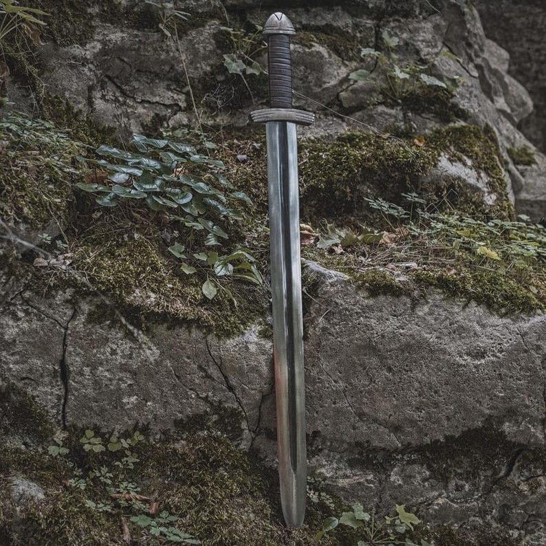 May include: A long, silver sword with a dark handle and crossguard is displayed against a moss-covered rock background. The sword's blade is polished, reflecting the light. The handle is wrapped in a dark material, and the pommel has a decorative design.