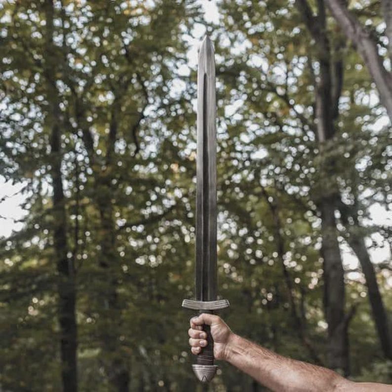 May include: A long, silver-colored sword with a dark brown handle is held up against a backdrop of green trees. The sword has a wide blade and a detailed hilt. The handle is wrapped in a dark material.