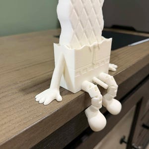 May include: A whimsical, white 3D-printed desk organizer shaped like a person. The design features a sponge-like top with a diamond pattern, a box-shaped body, and legs and feet dangling over the edge of a wooden desk.