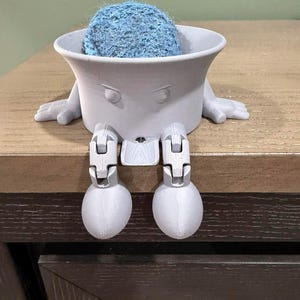 May include: A light gray, 3D-printed decorative container with a face, arms, and legs, holding a blue, fuzzy ball. The container is sitting on a wooden surface. The legs are bent, and the arms are resting on the surface.