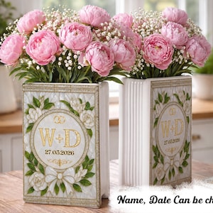May include: Two rectangular white ceramic vases featuring gold-trimmed borders and a central oval emblem with the gold letters W+D, two interlocking rings, and the date 27.05.2026, surrounded by painted white flowers, green leaves, and a white bow.