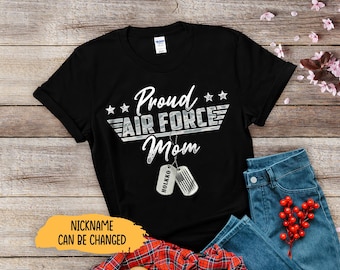 May include: Black t-shirt with the text "Proud Air Force Mom" in white script and a silver emblem. Includes two dog tags with the name "HOLKKO". The shirt is paired with blue jeans and a red berry sprig.