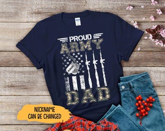 May include: Navy blue t-shirt with the text "PROUD ARMY DAD" in white and camouflage lettering. The design includes an American flag graphic and military-style dog tags. The shirt is paired with blue jeans and a red berry sprig.