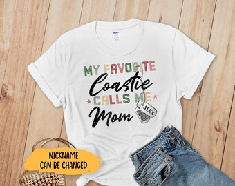 May include: White t-shirt with the text "MY FAVORITE Coastie CALLS ME Mom" in black script and colorful block letters. Includes dog tag pendants that say "ALEX". A pair of blue jeans is folded next to the shirt.