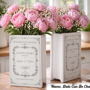May include: Two white ceramic book-shaped vases featuring a decorative gray border and the text "OUR GREATEST CHAPTER," "A LOVE STORY," "written by," "Sophia & Ethan," and "EST. 09.15.2026." The vases are filled with pink peonies and baby's breath.