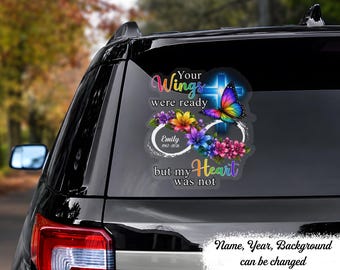 May include: A car window decal featuring a colorful butterfly, a blue glowing cross, and floral accents arranged around an infinity symbol. The text reads: Your Wings were ready but my Heart was not, with the name Emily and dates 1967-2026.
