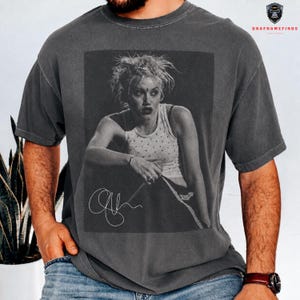 Peut inclure: T-shirt gris fonc&eacute; avec une image en noir et blanc d'une personne et une signature. L'image sur le t-shirt est un portrait d'une personne portant un d&eacute;bardeur. Le t-shirt a un col rond et des manches courtes.