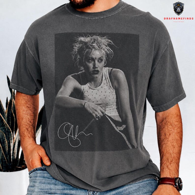 Peut inclure: T-shirt gris fonc&eacute; avec une image en noir et blanc d'une personne et une signature. L'image sur le t-shirt est un portrait d'une personne portant un d&eacute;bardeur. Le t-shirt a un col rond et des manches courtes.
