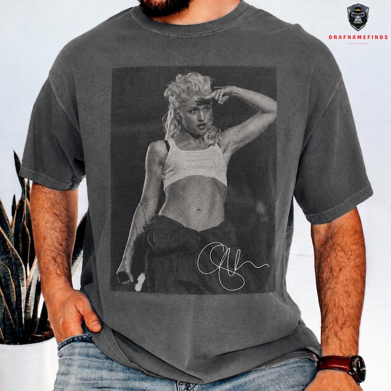 Peut inclure: T-shirt gris fonc&eacute; avec une image en noir et blanc d'une femme en crop top et salopette. L'image comprend une signature. Le t-shirt a un col rond et des manches courtes.