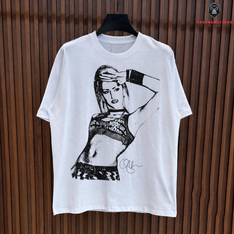 Peut inclure: T-shirt blanc avec une illustration en noir et blanc d'une femme. Le graphisme comprend le texte "READY NODOUR NODOU" et une signature. Le t-shirt est suspendu &agrave; un cintre noir sur un fond en bois.
