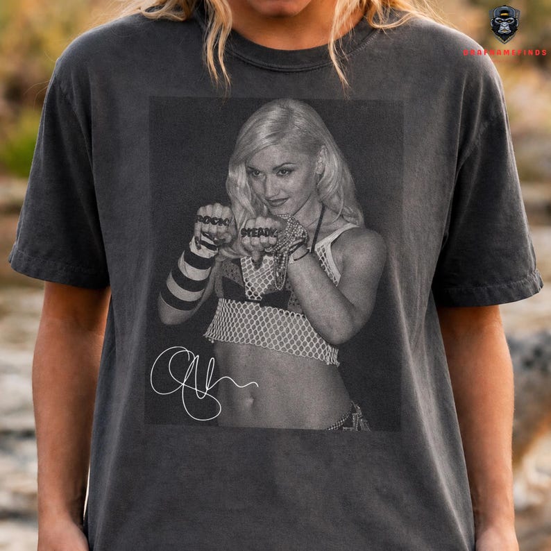 Peut inclure: T-shirt gris fonc&eacute; avec une image en noir et blanc d'une femme aux cheveux blonds. L'image comprend les mots "ROCK STEADY" sur les jointures et une signature. Le t-shirt est de style vintage.