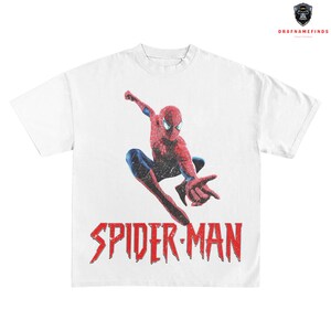 May include: White t-shirt featuring a distressed graphic of Spider-Man in a dynamic pose. The superhero is depicted in his classic red and blue suit, with the words "SPIDER-MAN" in bold red letters below.