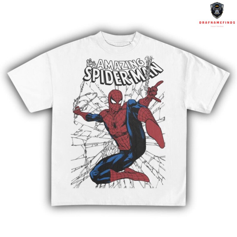 May include: White t-shirt featuring a graphic of Spider-Man in a dynamic pose, breaking through a web-like structure. The text "The Amazing Spider-Man" is printed above the character in black. The superhero is depicted in his classic red and blue suit.