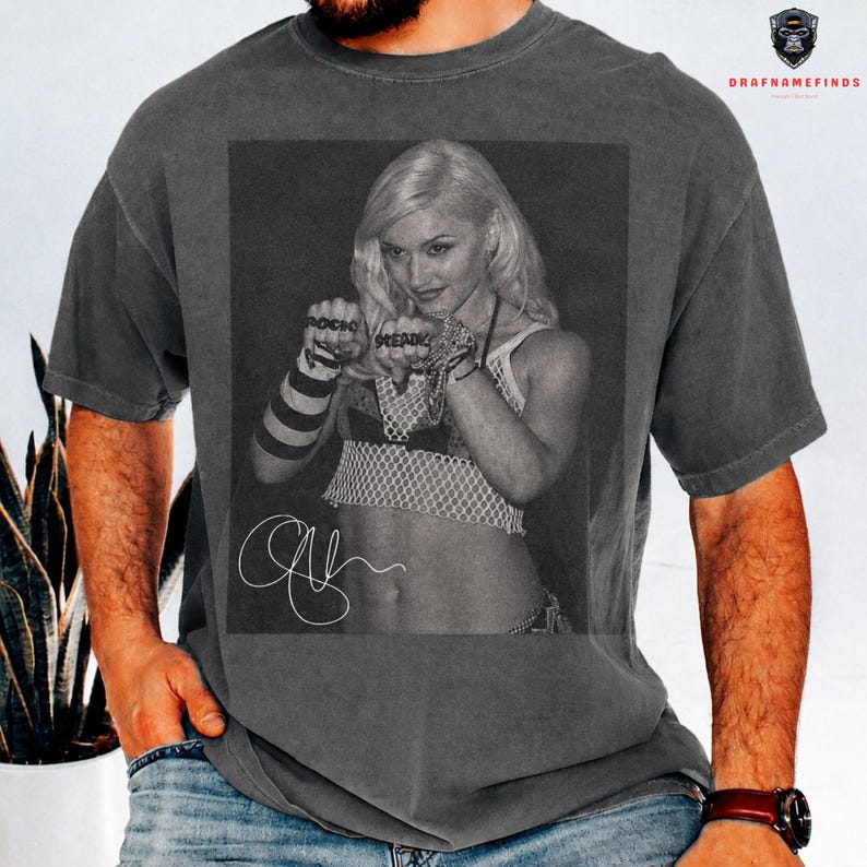 Peut inclure: T-shirt gris fonc&eacute; avec une image en noir et blanc d'une femme blonde. L'image comprend les mots "ROCK" et "BREATHE" sur les poings de la femme, ainsi qu'une signature. Le t-shirt a un aspect vintage.